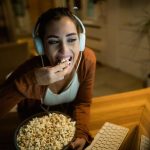 jovem mulher usando fones de ouvido enquanto come pipoca e assiste algo na internet em seu computador em casa 637285 4437