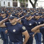 Policiais em uniforme fazem continência durante cerimônia pública em área urbana