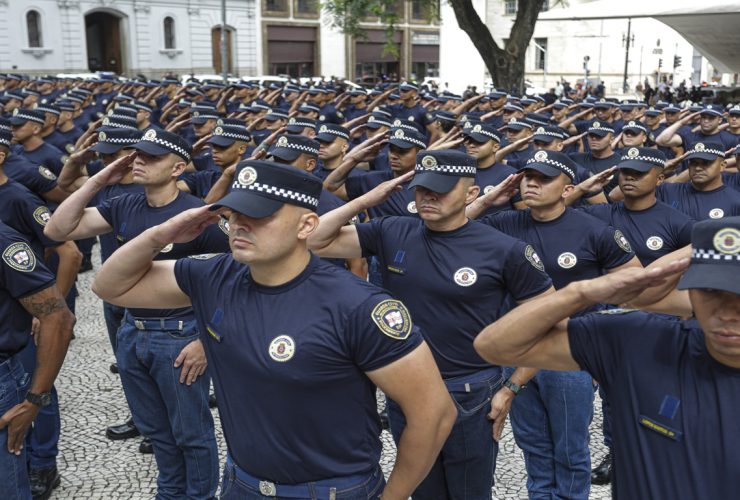 Policiais em uniforme fazem continência durante cerimônia pública em área urbana